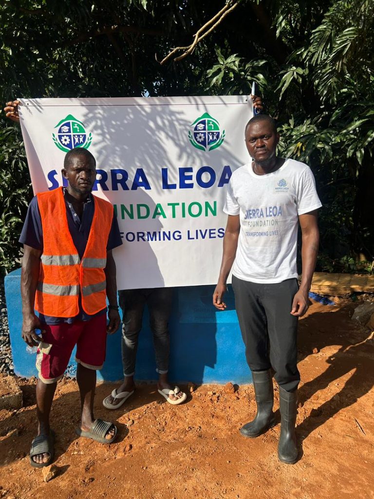 Two men stand in front of a banner reading “Sierra Leoa Foundation – Transforming Lives,” with trees and a blue water installation behind them. One man wears a bright orange safety vest, and the other wears a Sierra Leoa Foundation T-shirt and black boots.