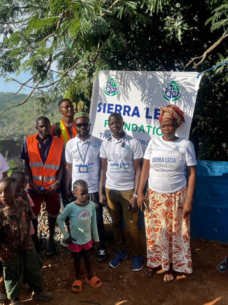 A group of adults and children stand outdoors in front of a “Sierra Leoa Foundation – Transforming Lives” banner. Several people wear Sierra Leoa Foundation T-shirts, while others, including children, stand beside them under the shade of a tree.
