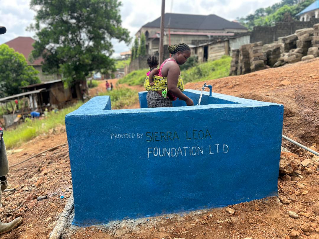 A woman and a young child collect clean running water from a newly built blue community tap station labeled “Provided by Sierra Leoa Foundation LTD,” located on a dirt hillside with homes and trees in the background.