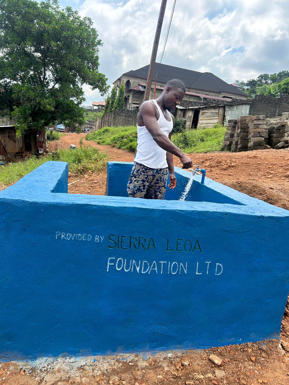 A young man fills a container with running water from a blue community tap system labeled “Provided by Sierra Leoa Foundation LTD,” set along a dirt road with houses, trees, and stone blocks in the background.