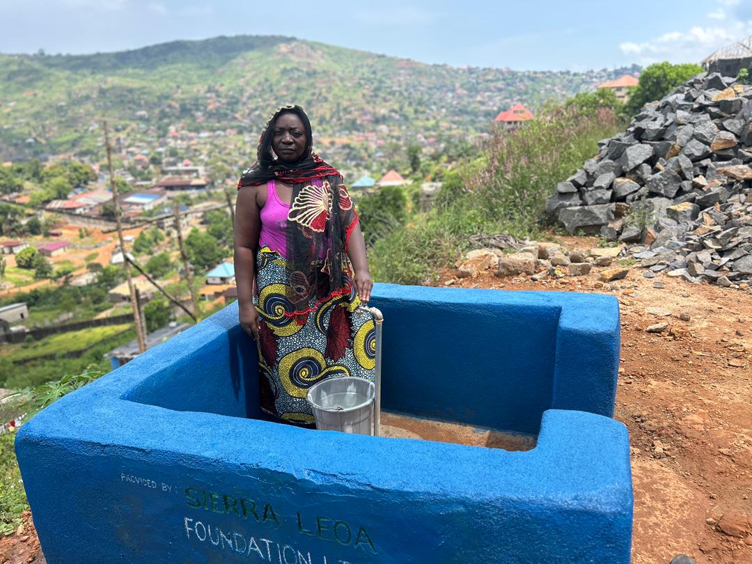A woman stands at a newly built blue water access point labeled “Sierra Leoa Foundation LTD,” with a metal bucket beside her. The background shows a hillside community with houses, greenery, and distant mountains.