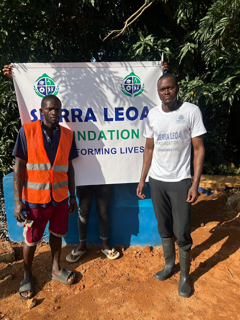 Two men stand in front of a banner reading “Sierra Leoa Foundation – Transforming Lives,” with trees and a blue water installation behind them. One man wears a bright orange safety vest, and the other wears a Sierra Leoa Foundation T-shirt and black boots.